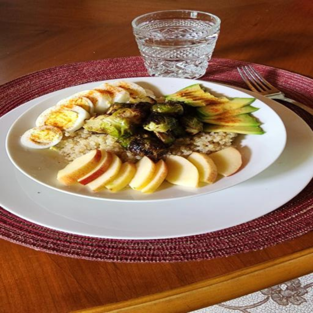 A bowl and a glass of water. The bowl contains quinoa, roasted brussel sprouts, sliced avocado, a sliced red apple, and a sliced boiled egg.